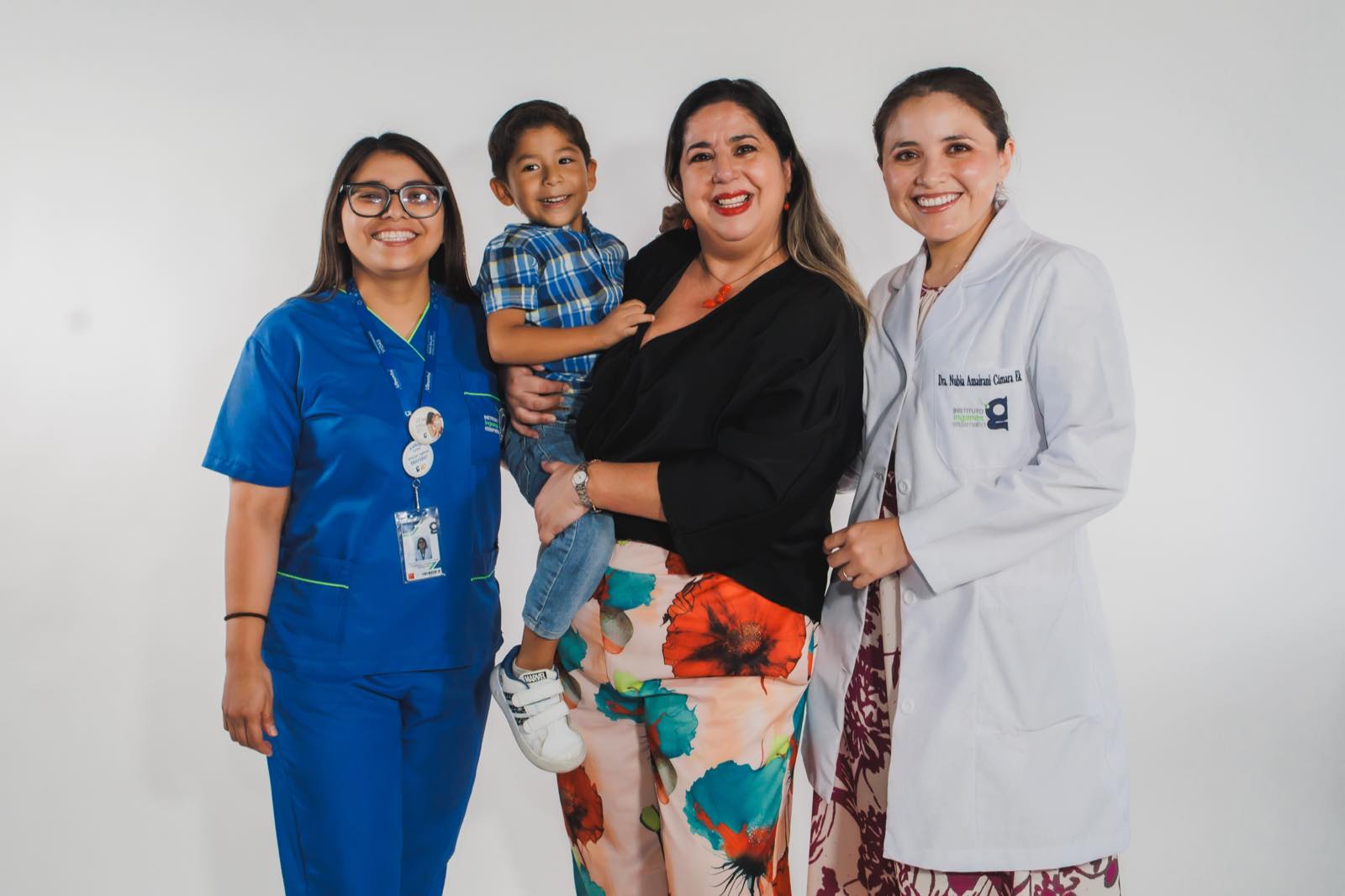 Doctora de Ingenes con bata blanca junto a una enfermera y una mamá que carga a su hijo