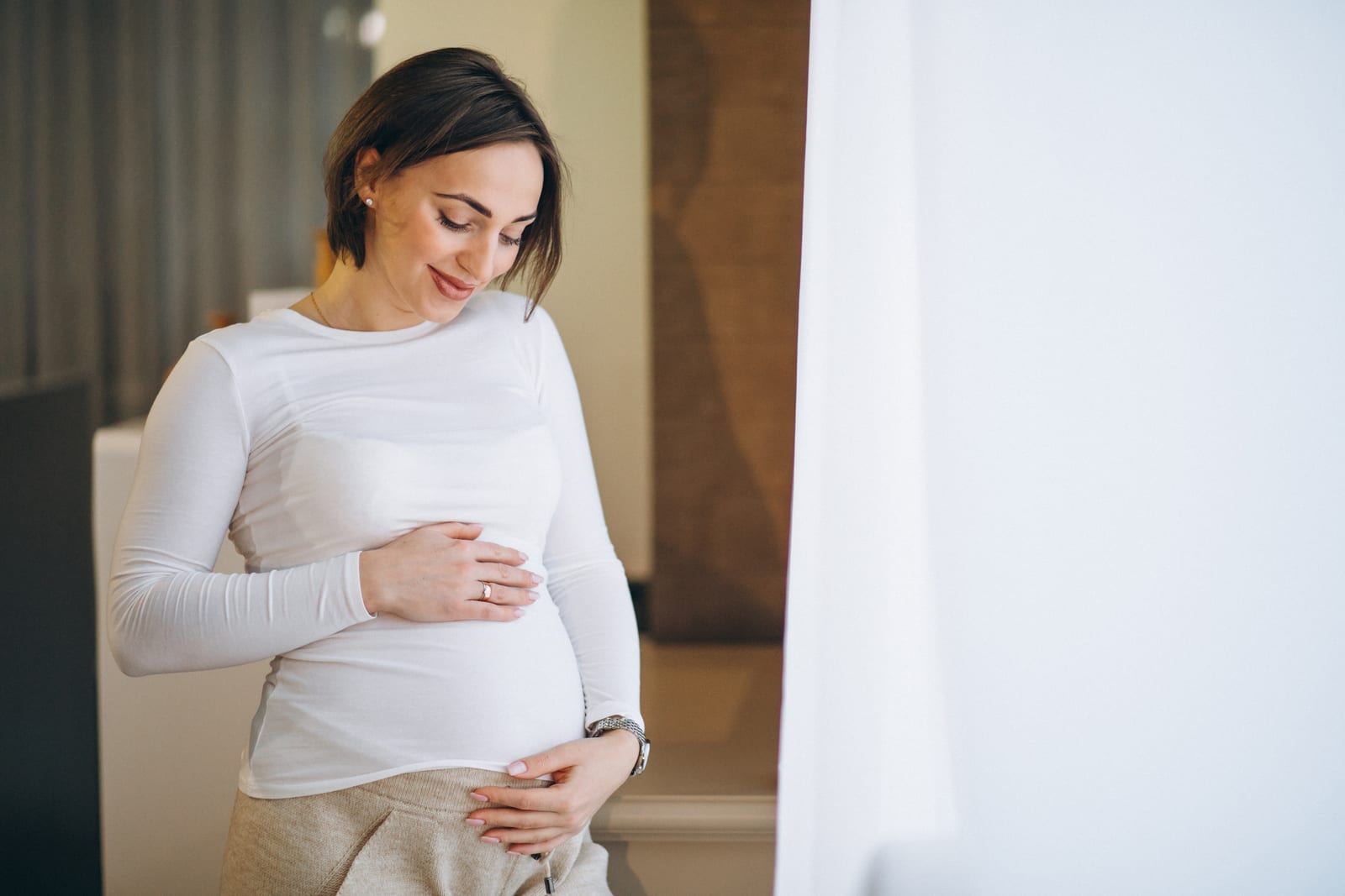 Pregnant woman smiling while looking at her belly by a window